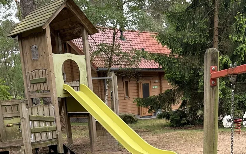 Jouw nieuwe logeerplek. | Fay zorglogeerhuis | Houten speeltoestel met een gele glijbaan bij de blokhut, omringd door bomen.